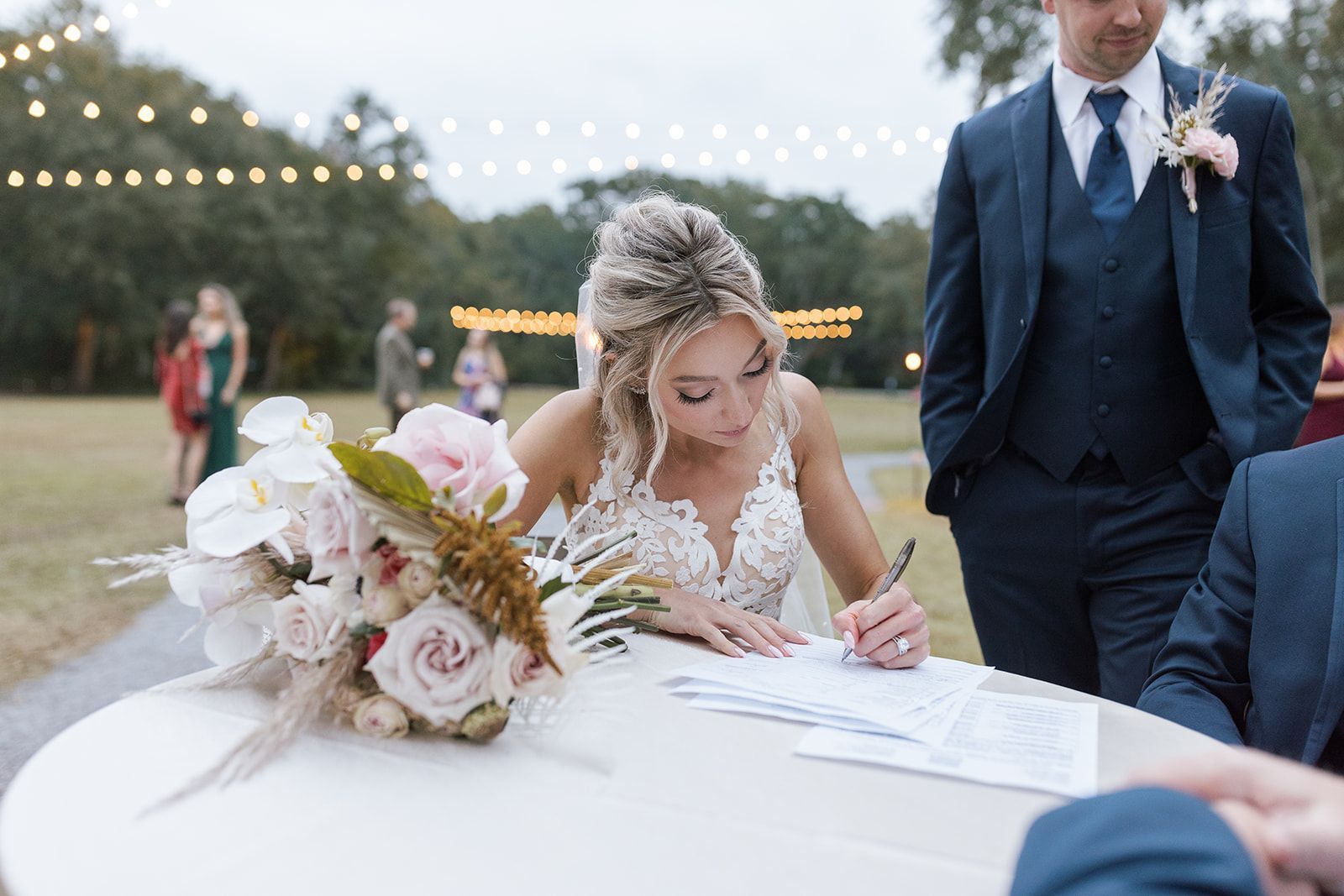 Bride signs a wedding document beside a bouquet at an outdoor reception table