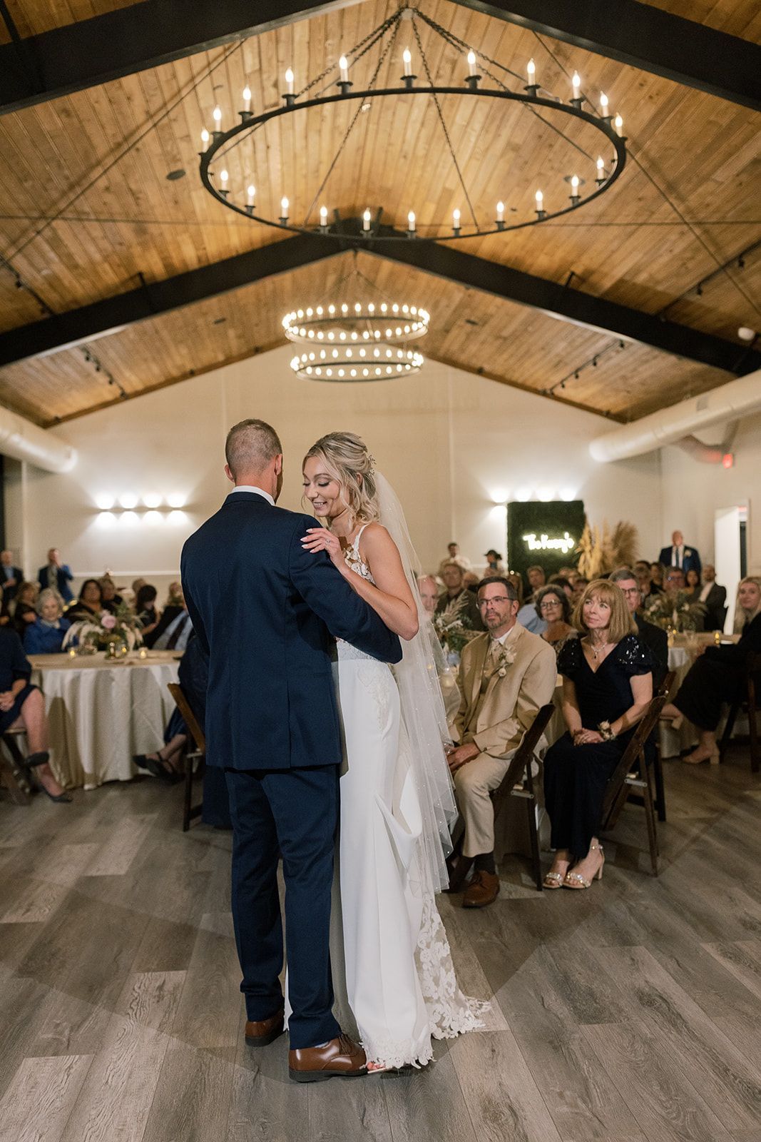 Wedding couple dancing under chandeliers in a rustic reception hall, guests watching nearby.