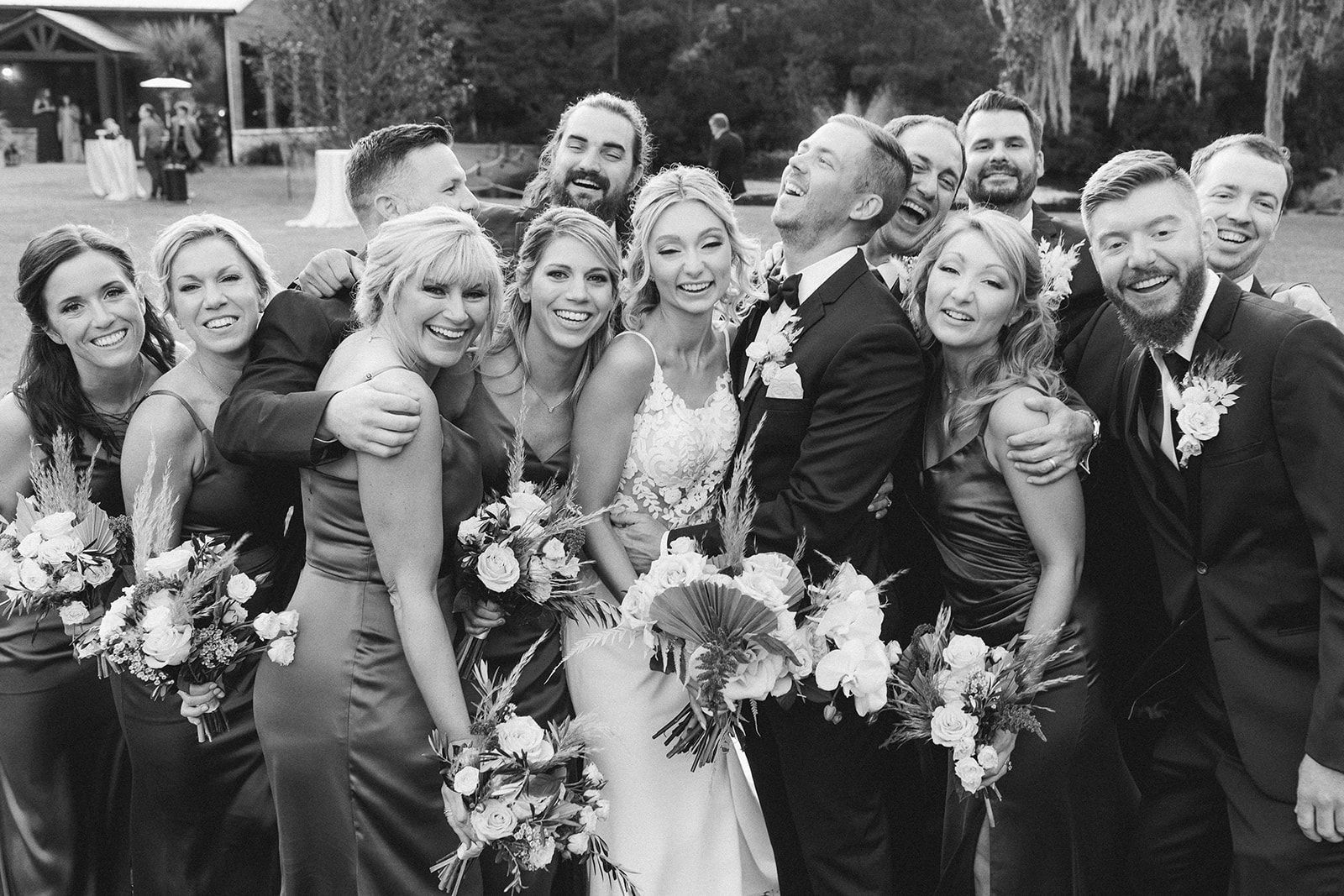 A group of wedding guests pose outdoors, smiling and holding bouquets in formal attire.