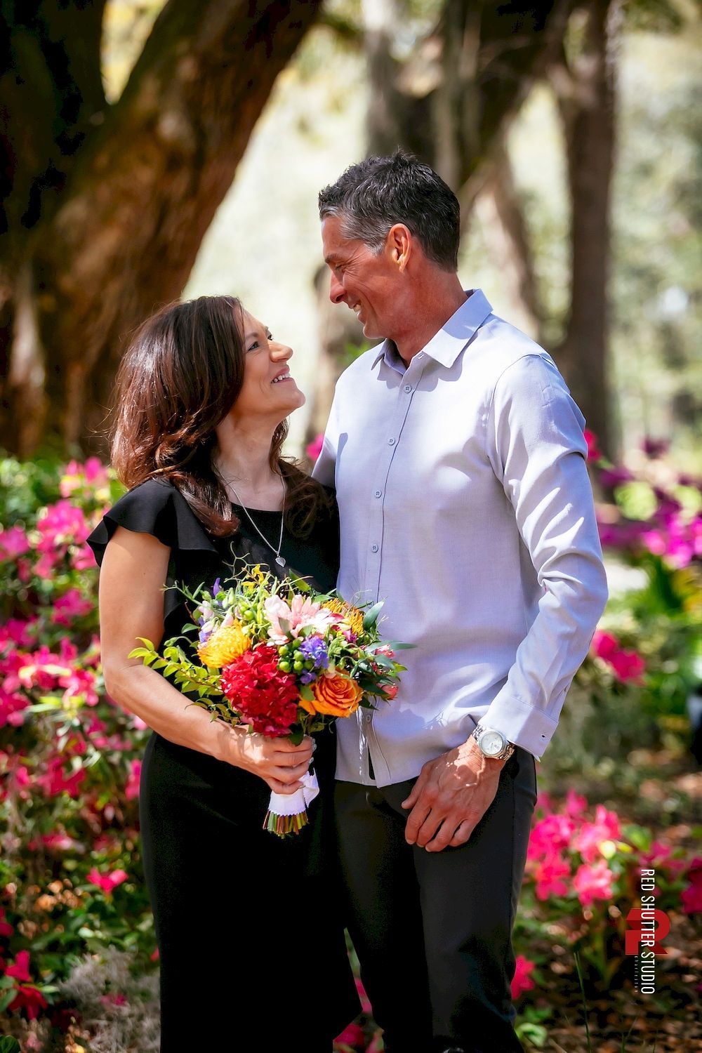 Couple smiling and holding flowers in a garden with pink blooms and trees