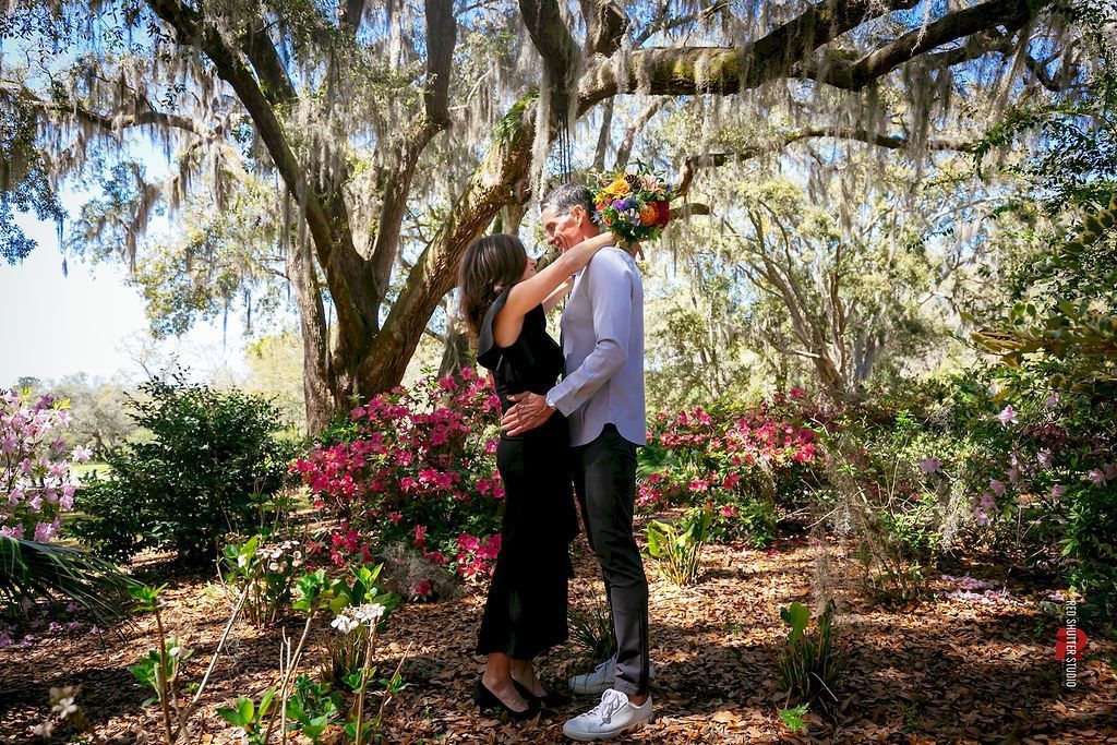Couple embracing in a flowering garden under trees, with sunlight and pink blossoms.