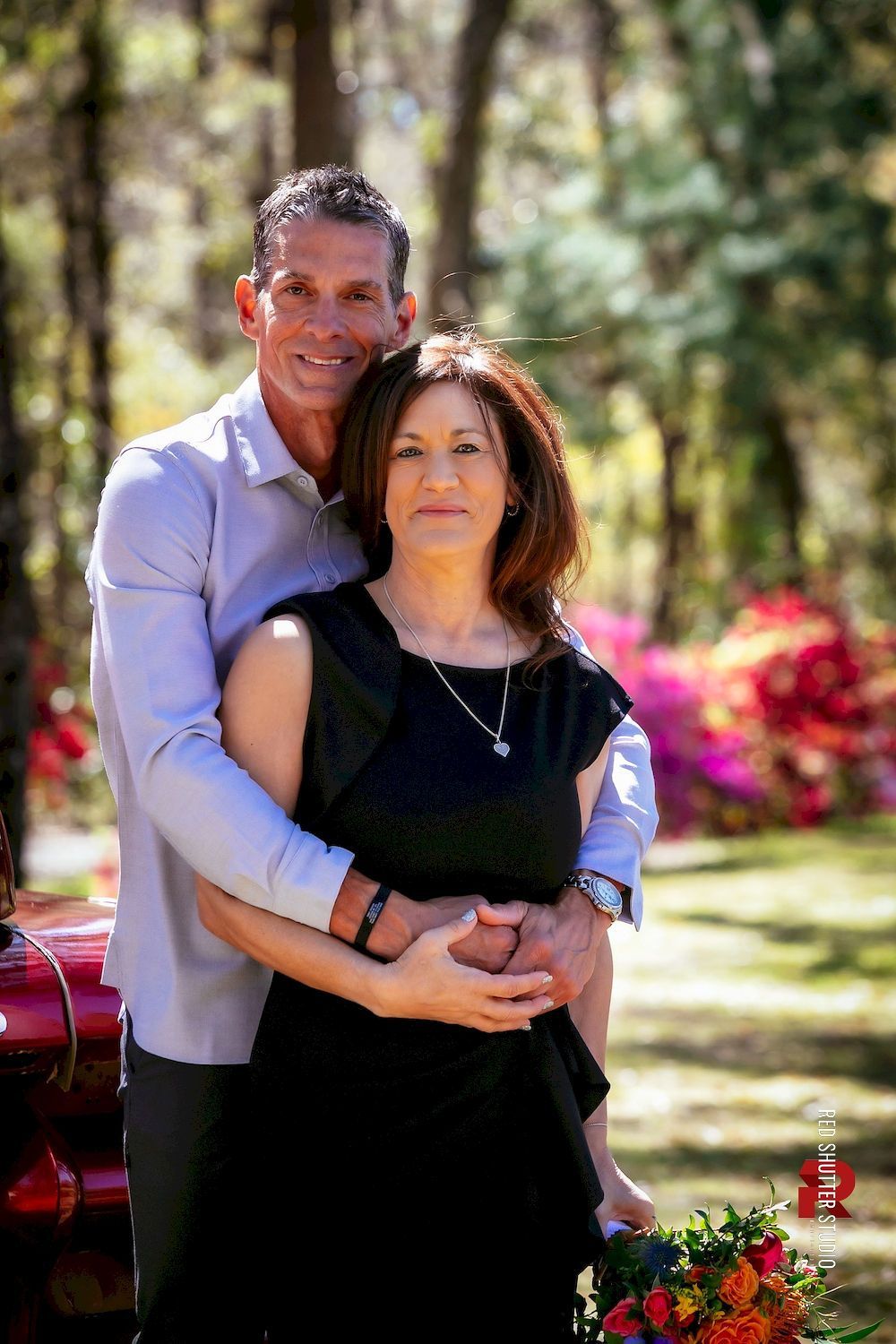 A couple embracing outdoors, with flowers and trees in the background.