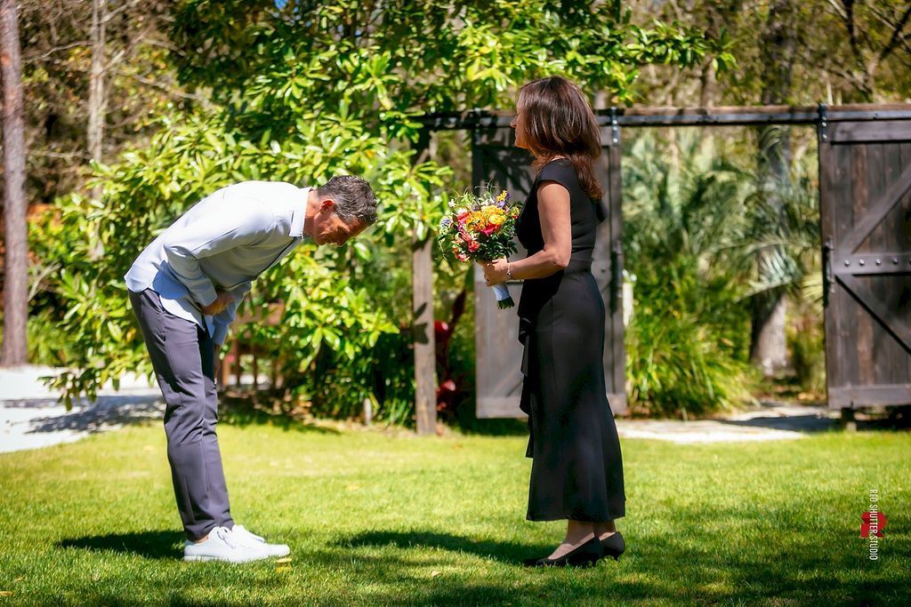 Two people standing on a lawn, one bending forward as the other holds a bouquet near a garden gate.