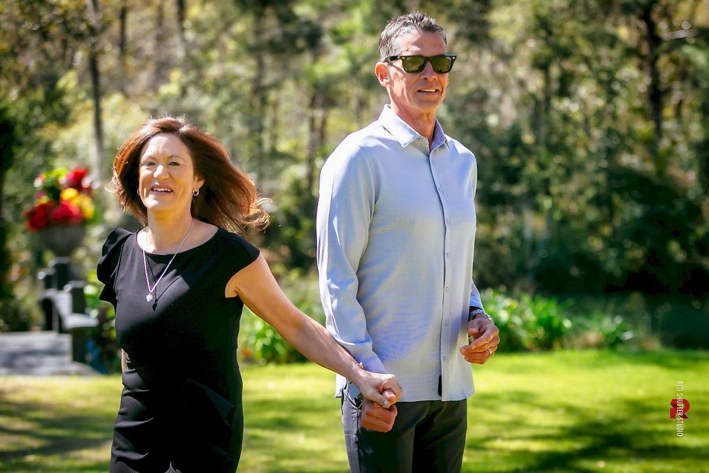 Two people holding hands and walking through a sunny garden, smiling.
