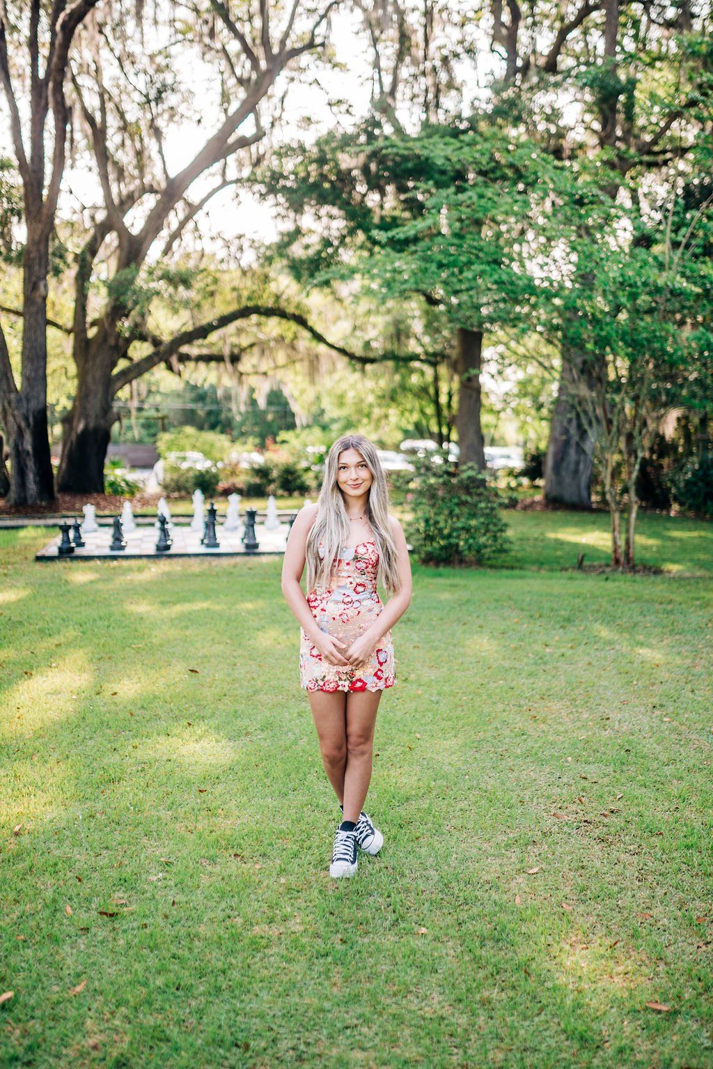 Woman in a floral dress standing on a grassy lawn in a park with trees and chessboard in the background