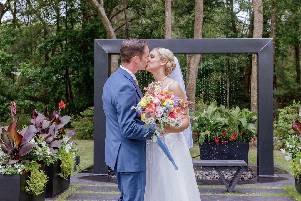 Bride and groom kiss in a garden wedding ceremony, holding a colorful bouquet under a black arch.