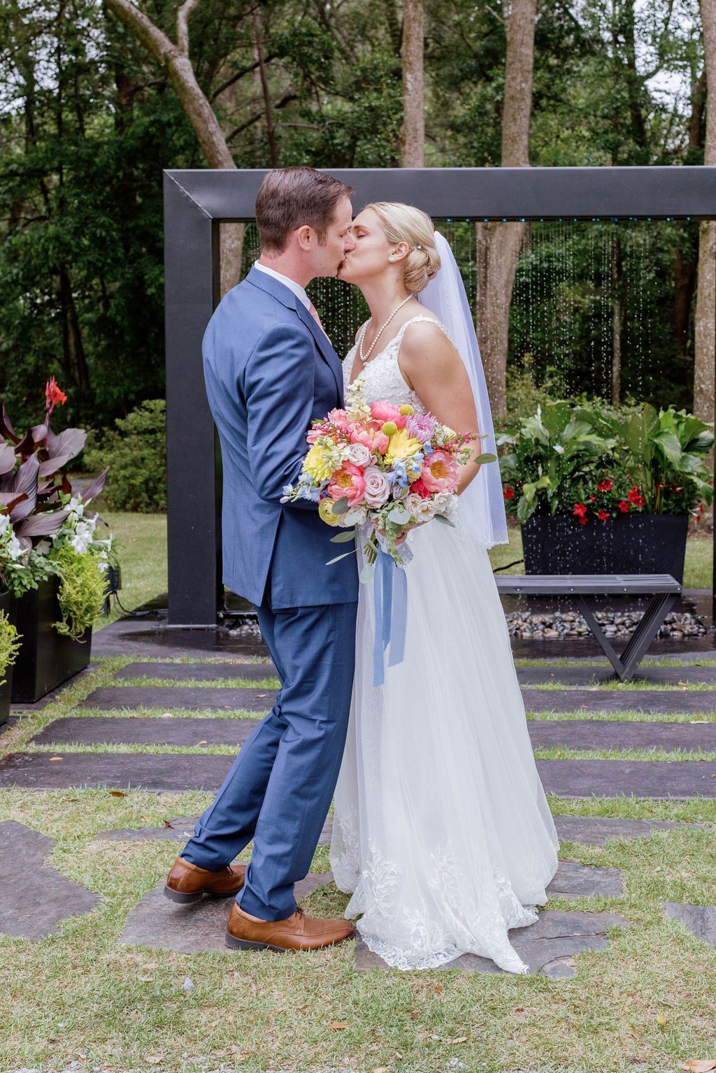 Wedding couple kissing under a dark wooden arch in a garden, bride holding a colorful bouquet