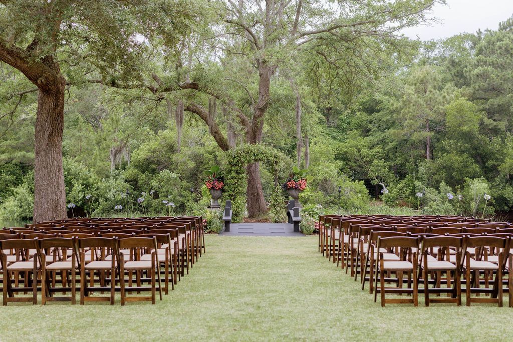 Outdoor wedding setup with rows of wooden chairs on a grassy aisle beneath large trees