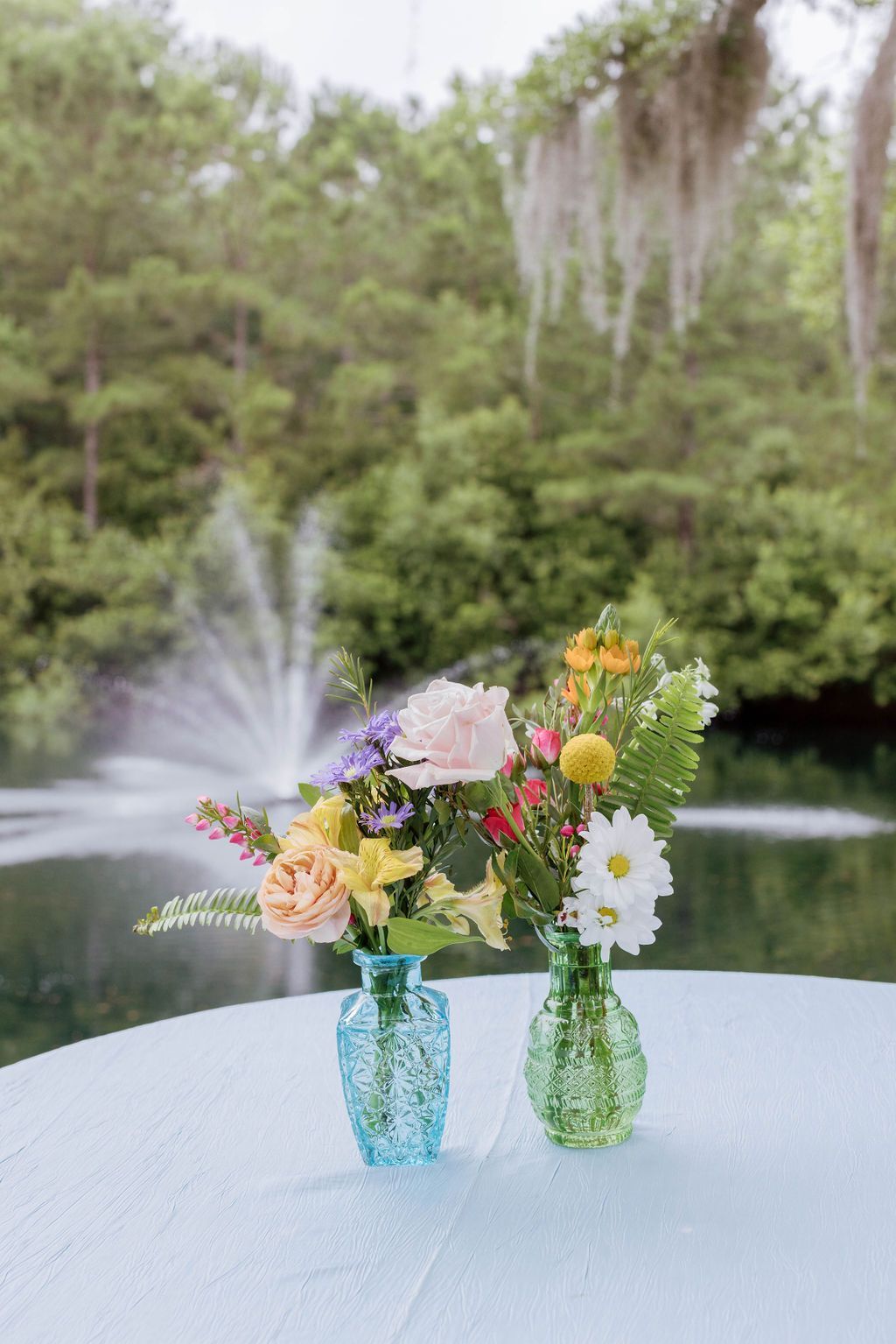 Two flower arrangements in blue and green vases on a table by a pond fountain.