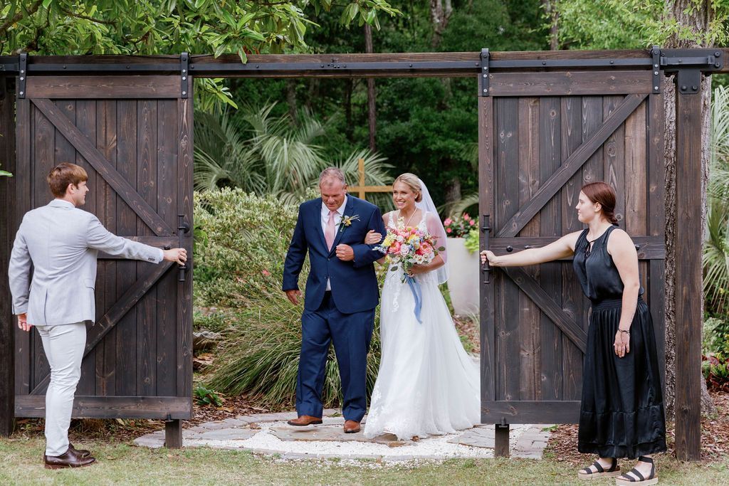 Wedding couple entering through open wooden barn doors, flanked by attendants in formalwear