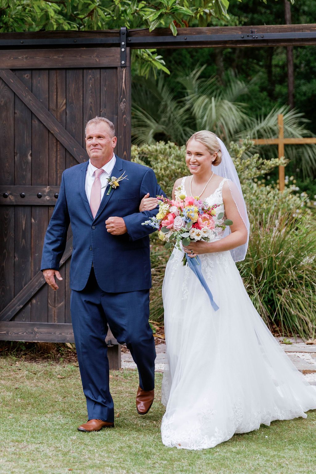Bride in white dress and groom in navy suit walking outdoors, holding hands and a bouquet