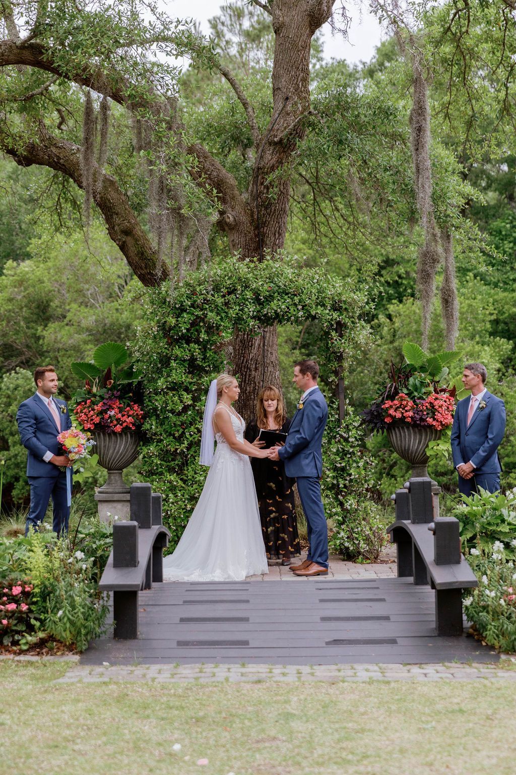 Bride and groom exchange vows on a garden stage under a large tree, flanked by floral arrangements.
