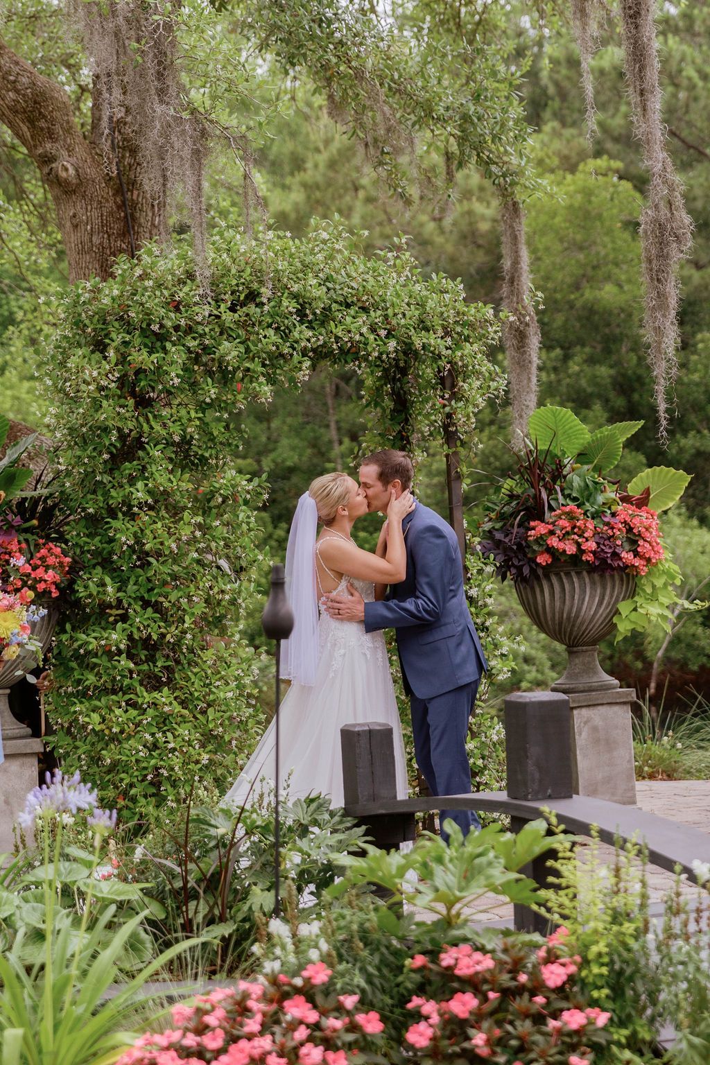 Bride and groom kissing in a lush garden beside flowers and tall trees