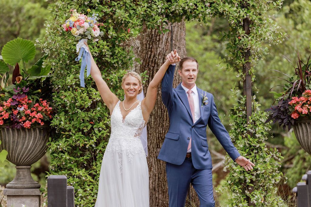 Newlyweds in blue and white outfits holding hands under a floral arch, smiling outdoors