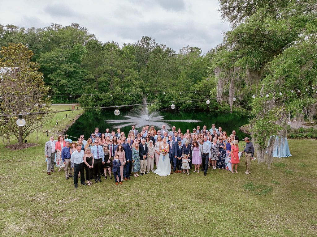 Large group posing on a grassy lawn by a lake, with fountains and trees in the background.