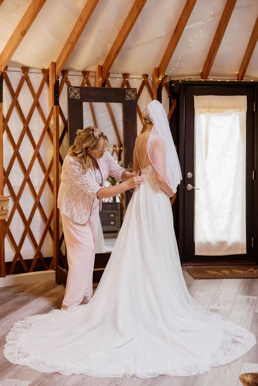 Bride in white gown with long train being adjusted by another woman inside a wooden yurt room
