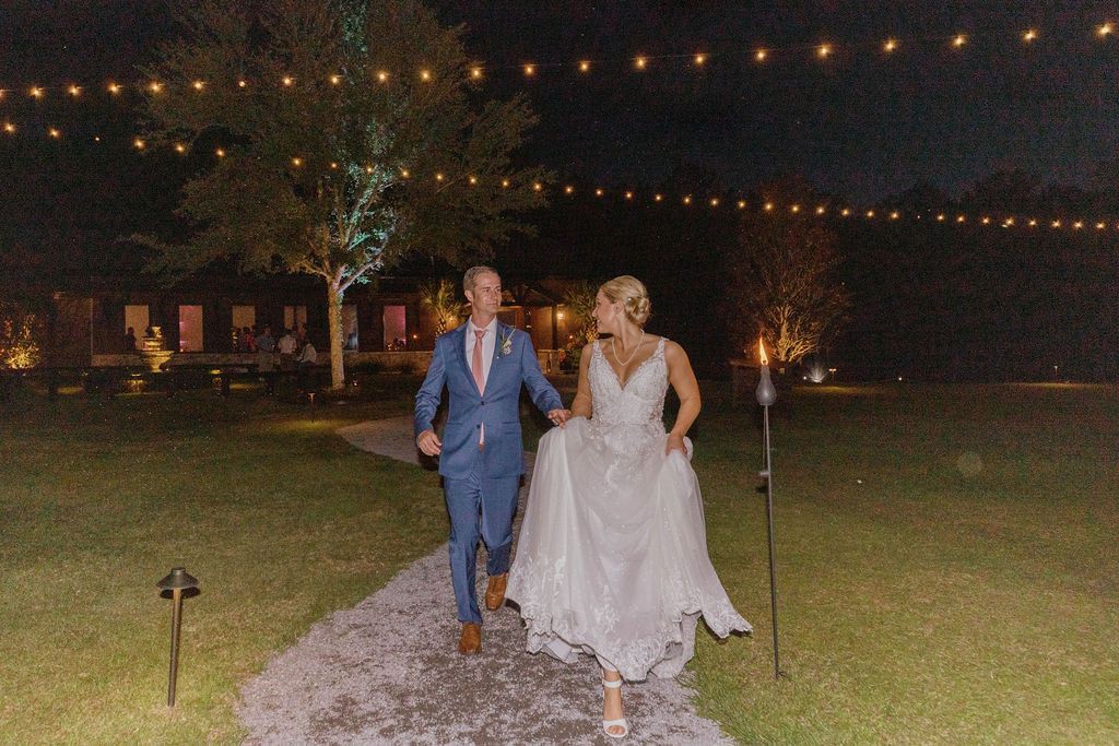Couple walking down a lit garden path at night, the bride in a white dress and the groom in a blue suit.