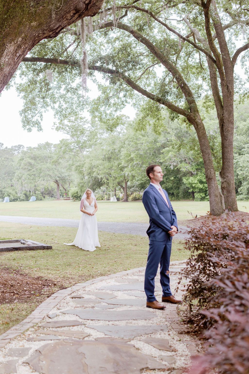 Man in a blue suit standing on a stone path under large trees, with a woman in white in the background.