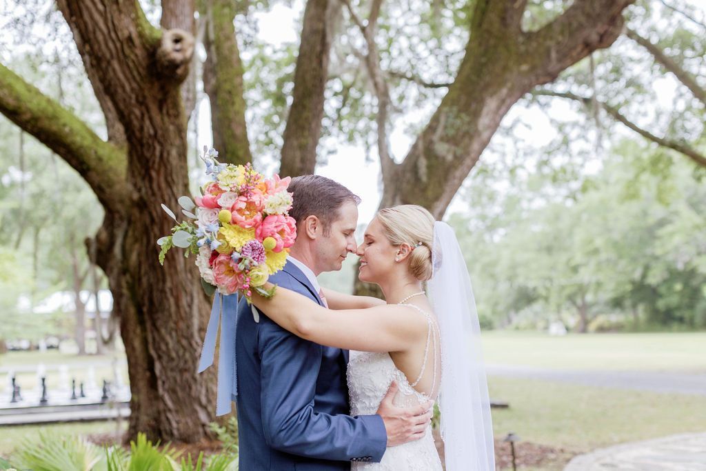 Bride and groom embracing under a large tree, with a colorful bouquet and veil in a park setting