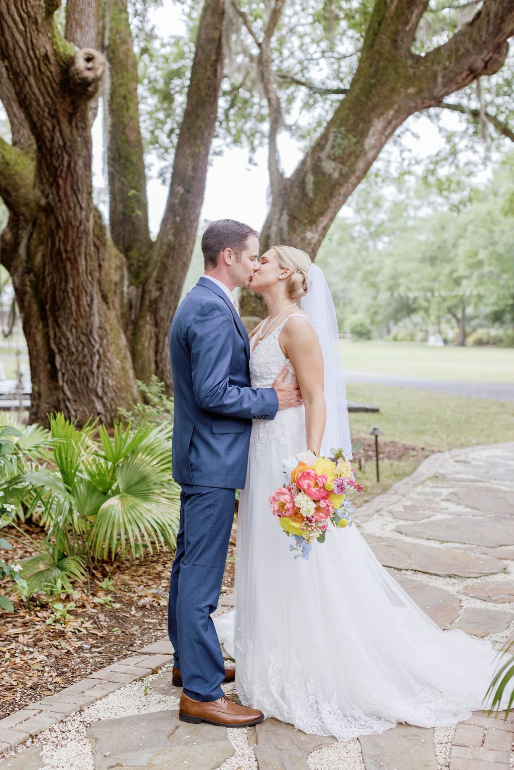 Wedding couple kissing outdoors by a tree, bride holding a colorful bouquet in a white gown, groom in a blue suit