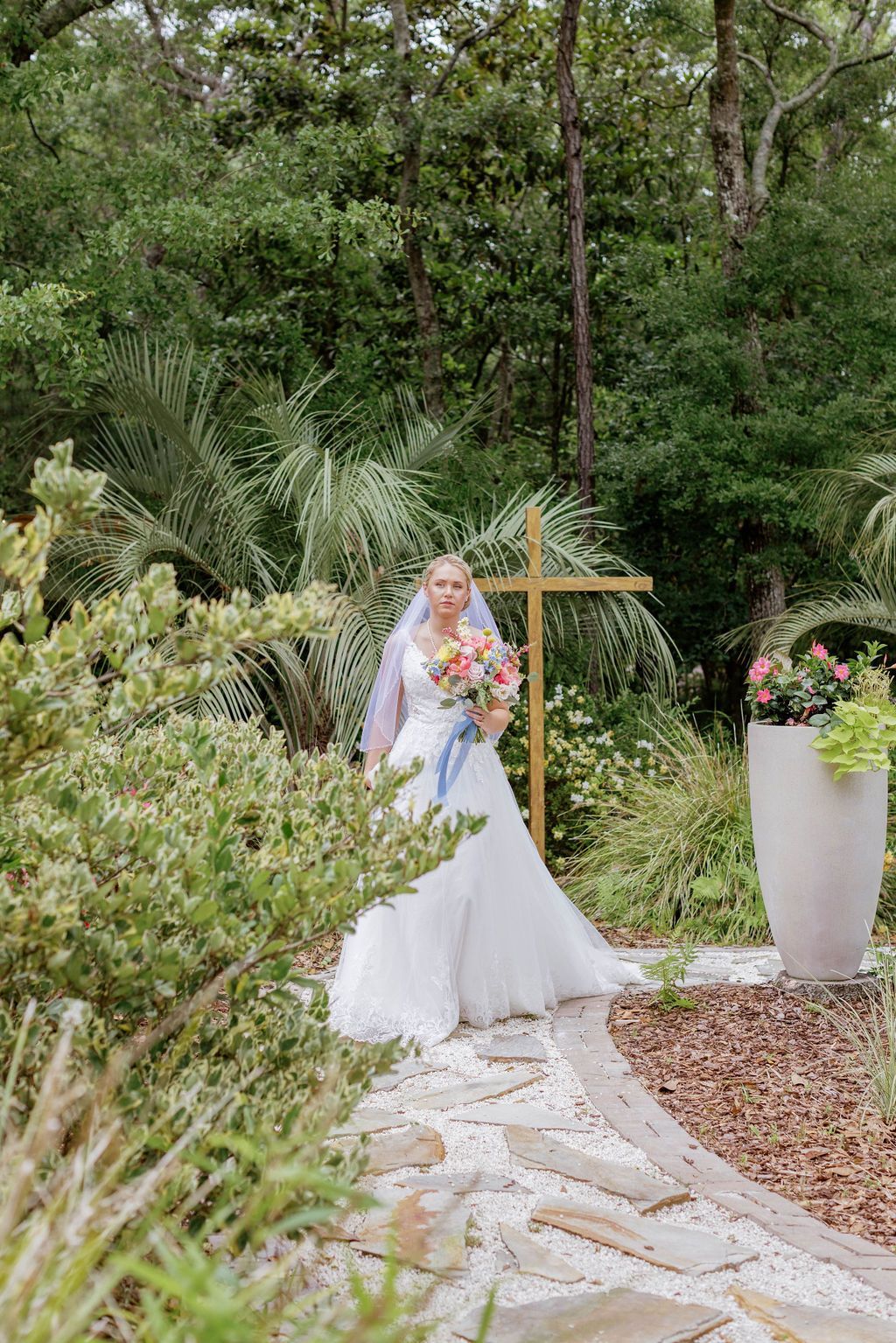 Bride in white gown holding bouquet beside a wooden cross in a lush garden path