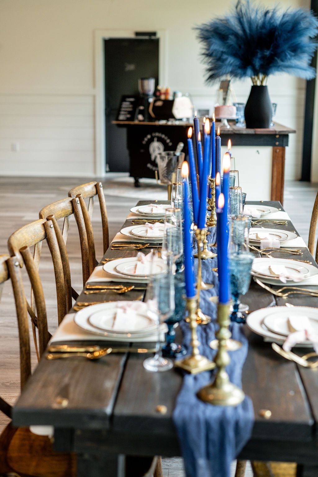 Long dining table set with white plates, gold candlesticks, blue accents, and a navy centerpiece in a bright room