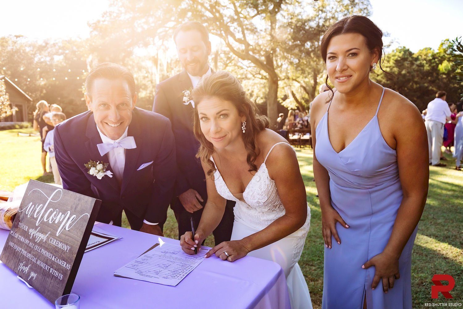 Three people signing a guest book at an outdoor wedding reception table in warm sunlight