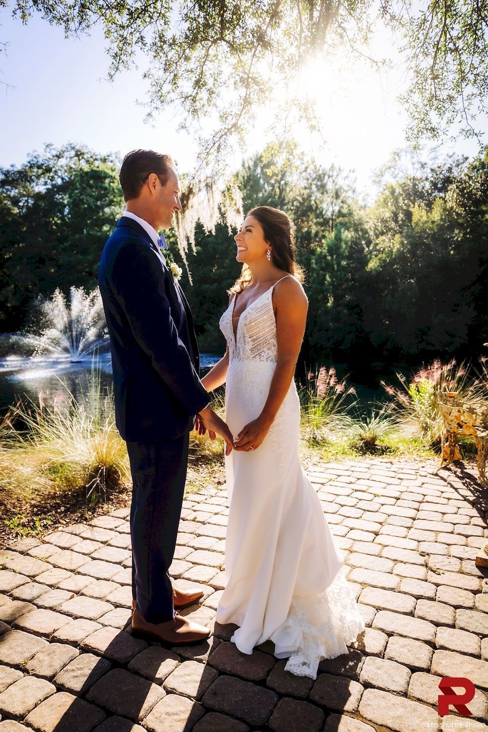 Couple in wedding attire holding hands on a sunlit cobblestone path in a garden