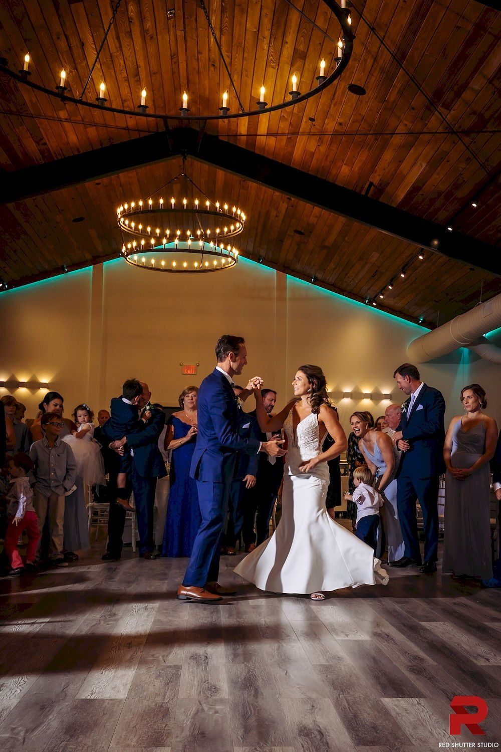 Bride and groom dancing at a rustic indoor wedding reception, guests gathered around them