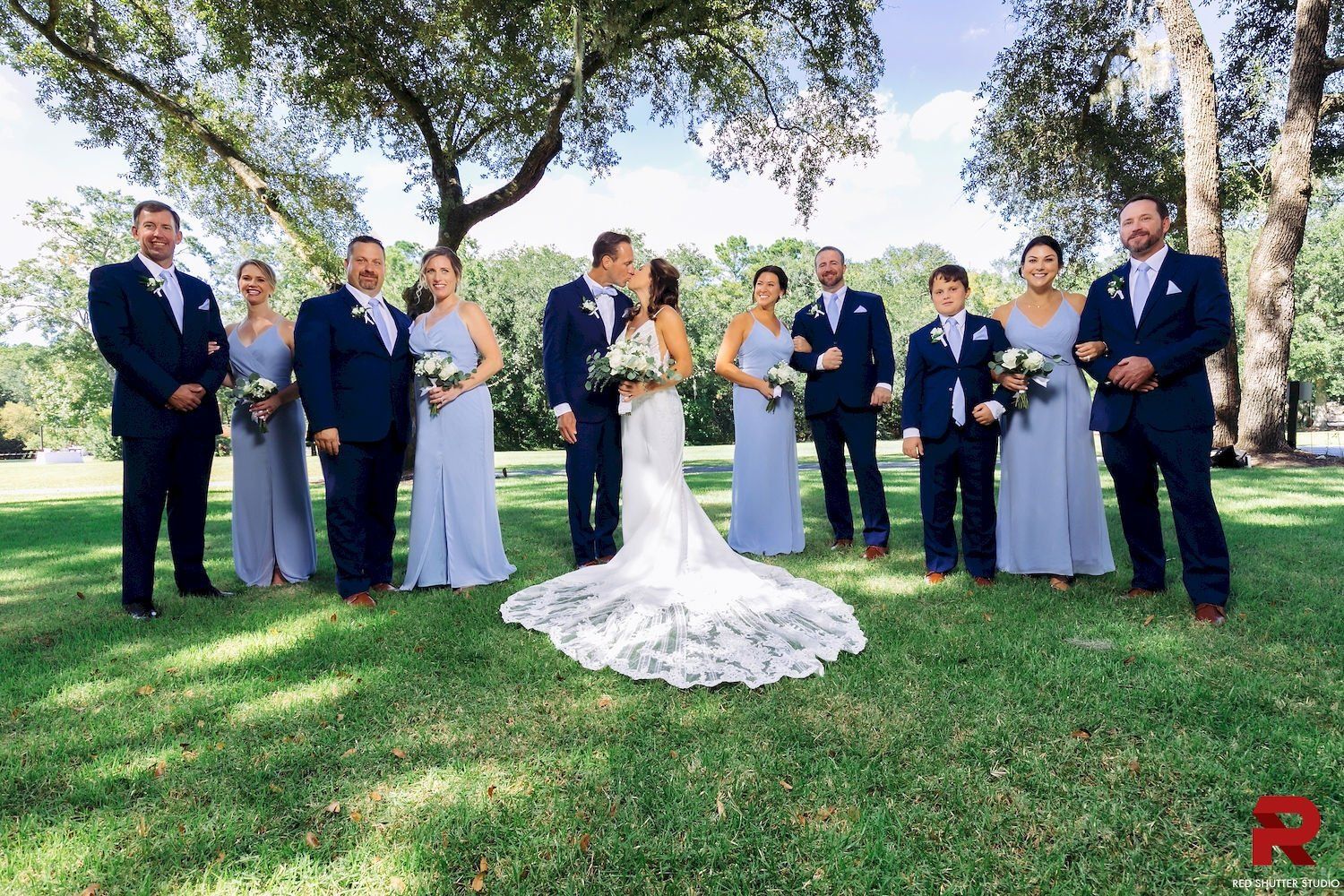 Wedding party posing outdoors on a sunny lawn, with the bride in a white gown and attendants in navy and lavender.