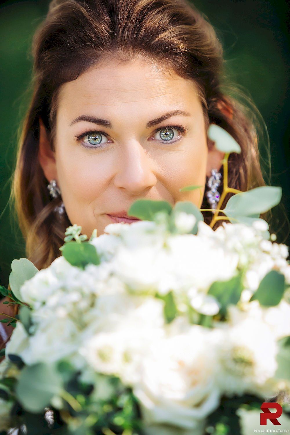 Woman peeking over a bouquet of white flowers, with bright blue eyes and a blurred green background