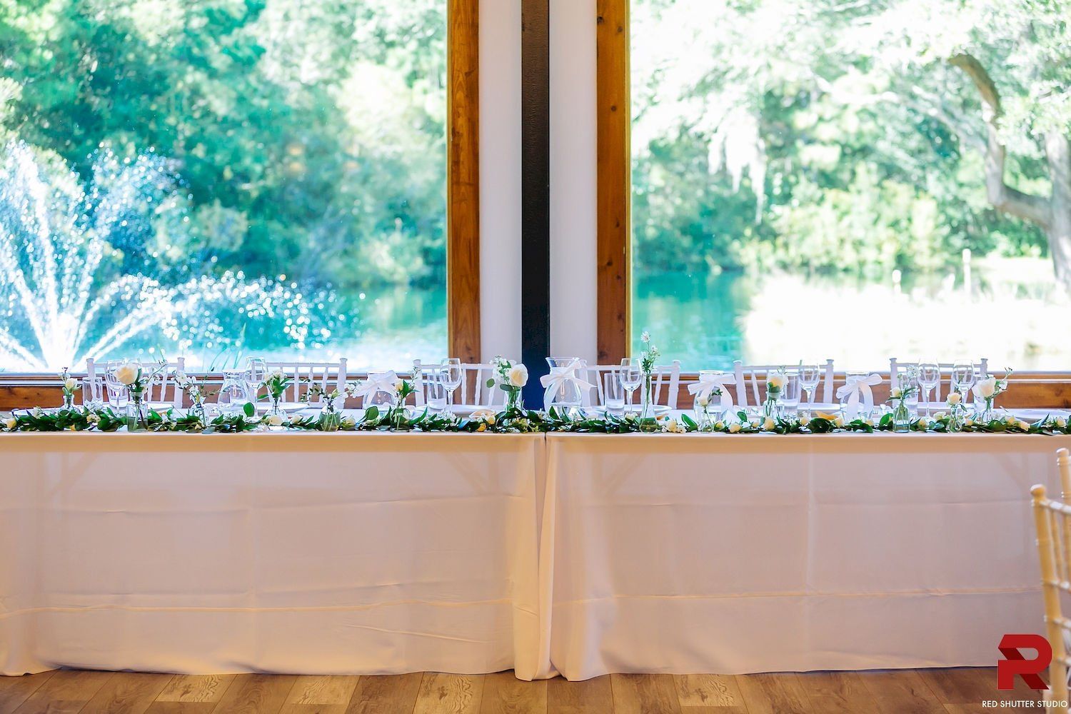 White-clothed banquet table beside large windows with floral arrangements and green trees outside.