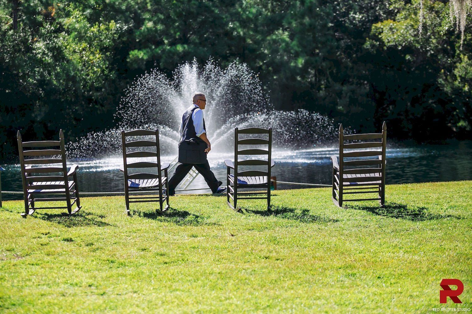 Person walking past chairs in a sunny park with a fountain spraying water in the background