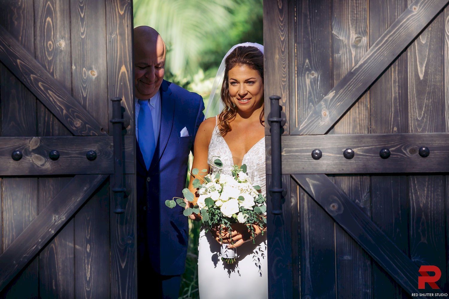Bride and groom peeking through open barn doors, bride holding white flowers and smiling.