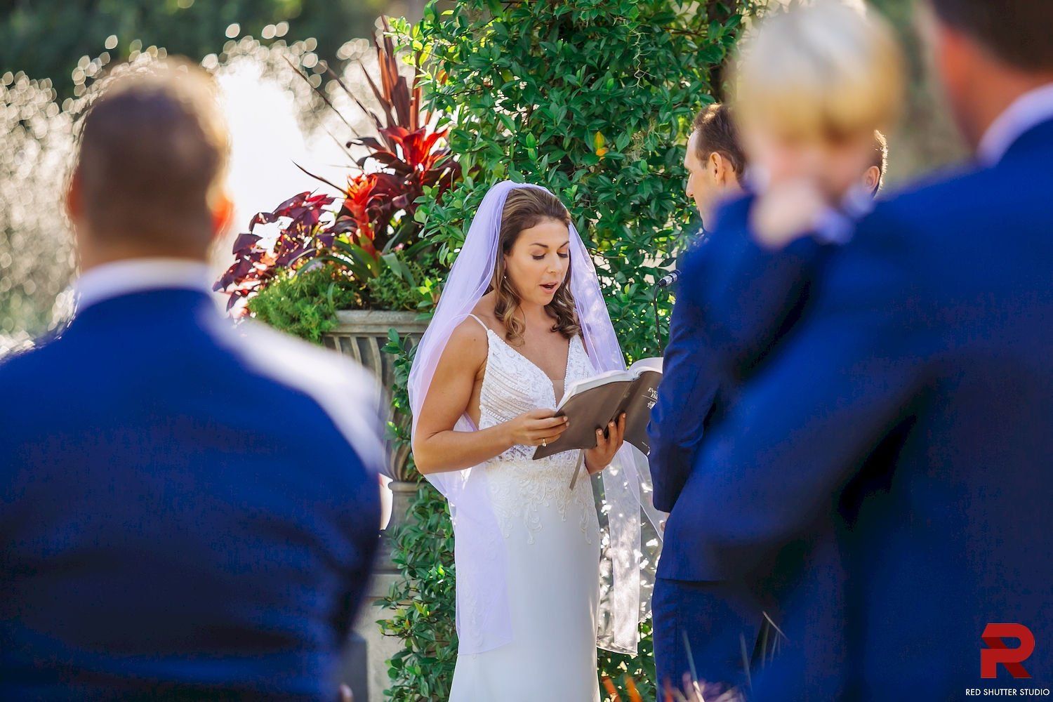 Bride in a white dress reading vows at an outdoor wedding ceremony, framed by guests in blue suits.