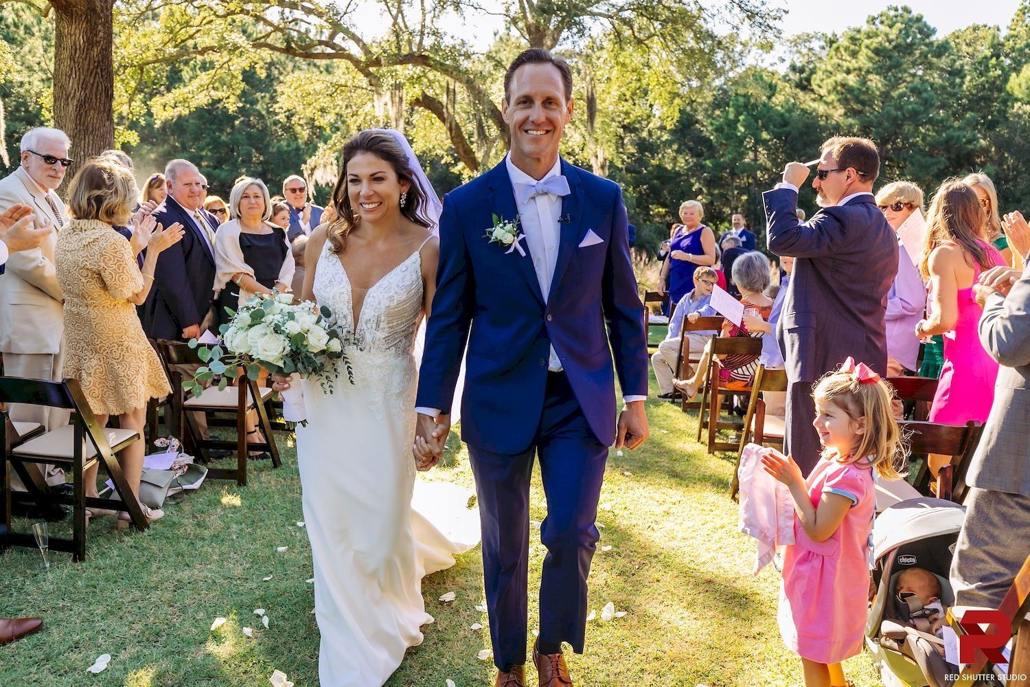 Wedding couple walking down outdoor aisle, bride in white dress and groom in blue suit, guests applauding