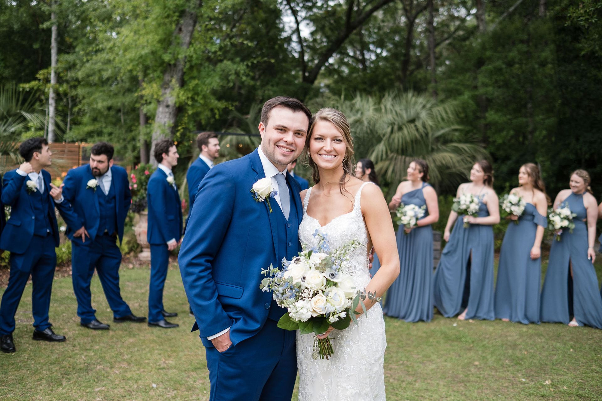 Bride and groom pose outdoors with wedding party in blue and gray dresses and suits behind them.