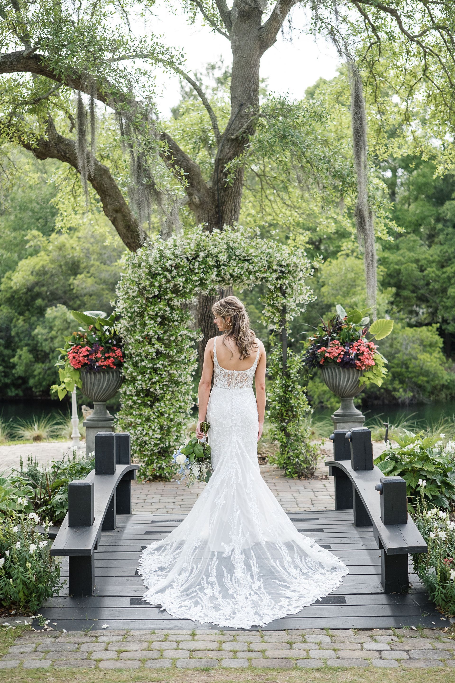 Bride in a white lace gown standing on a garden aisle with flowers and trees