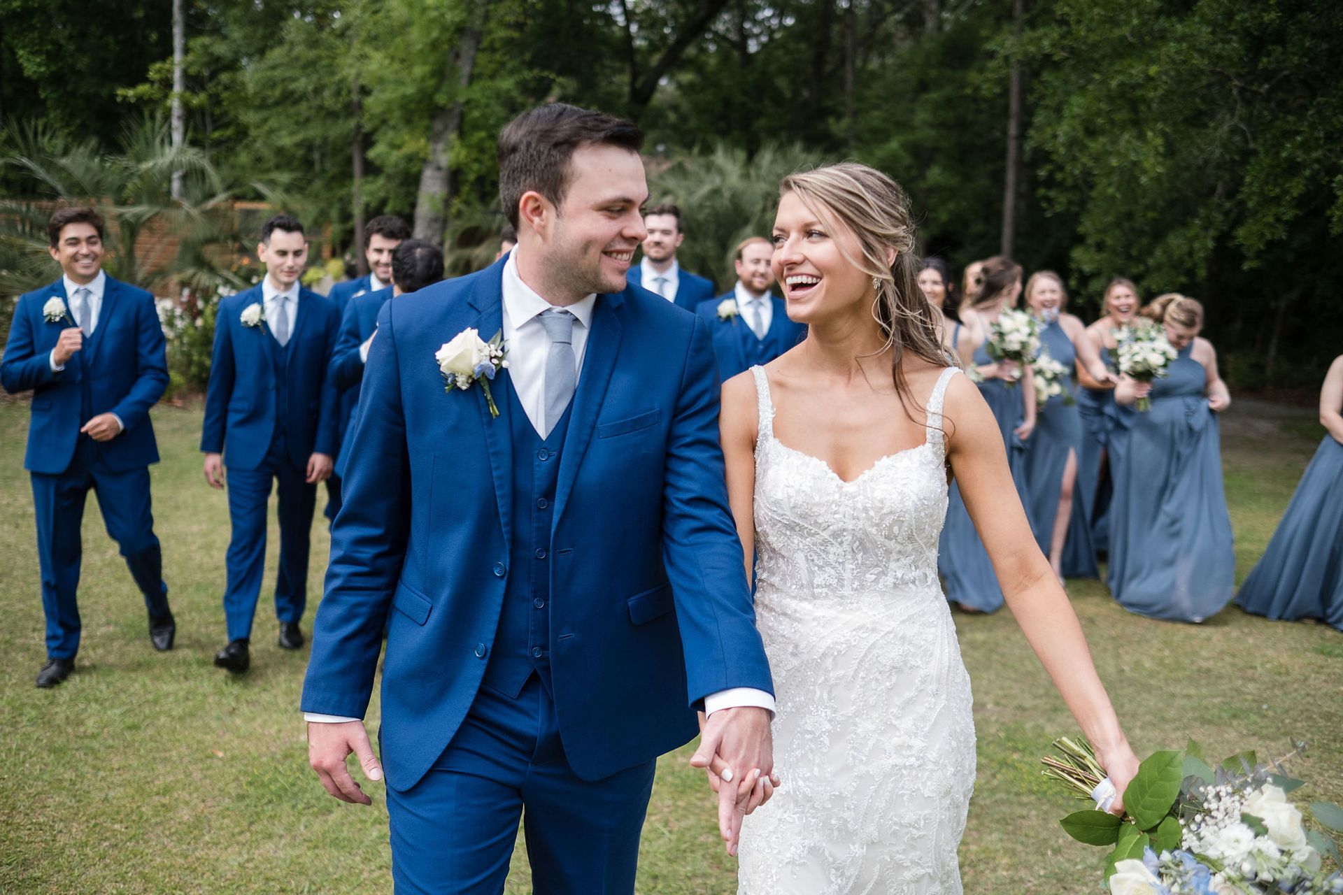 Newlywed couple in blue suit and white lace dress walking hand in hand outdoors, smiling at each other