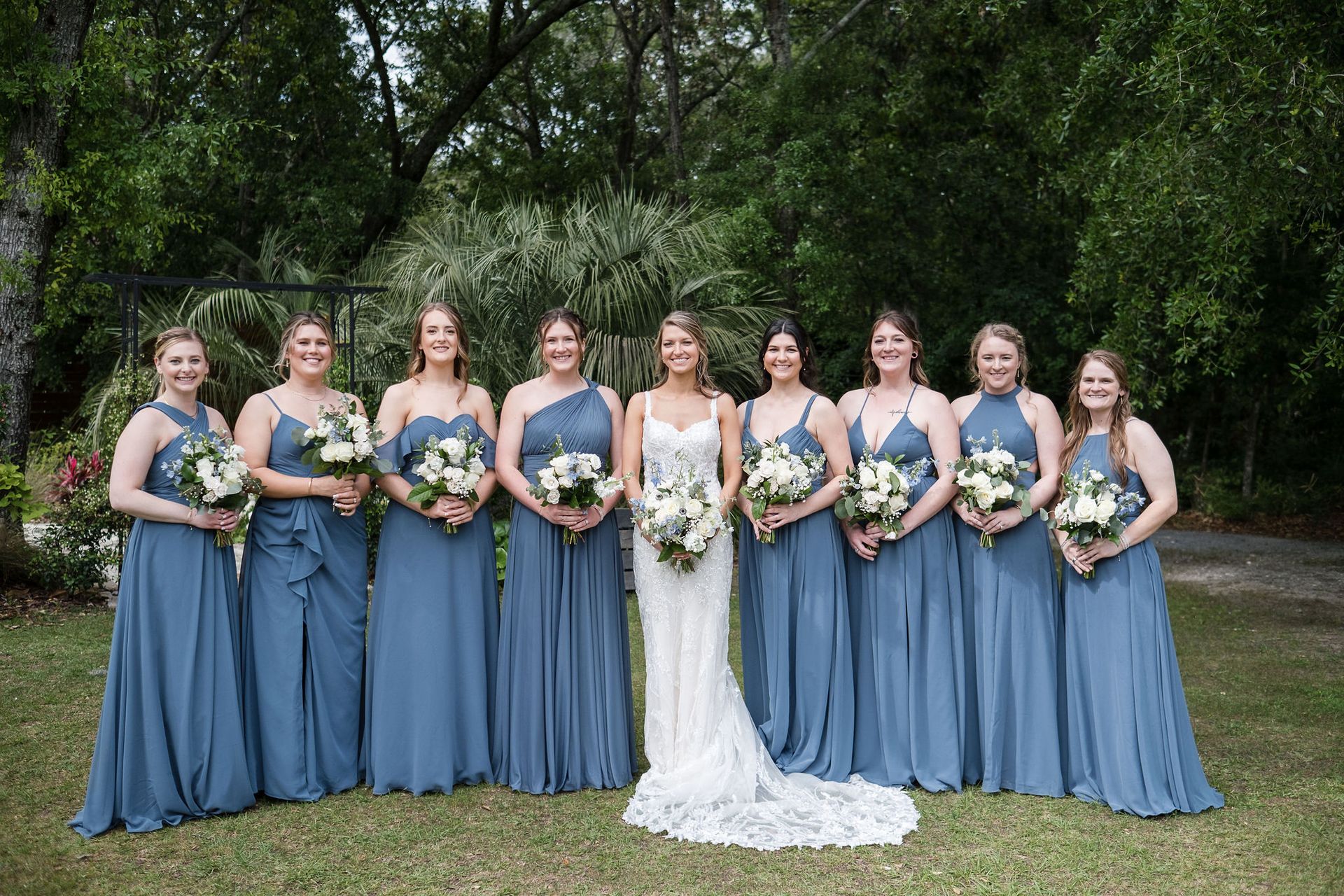Bridesmaids in blue dresses stand with a bride in a white gown holding bouquets outdoors