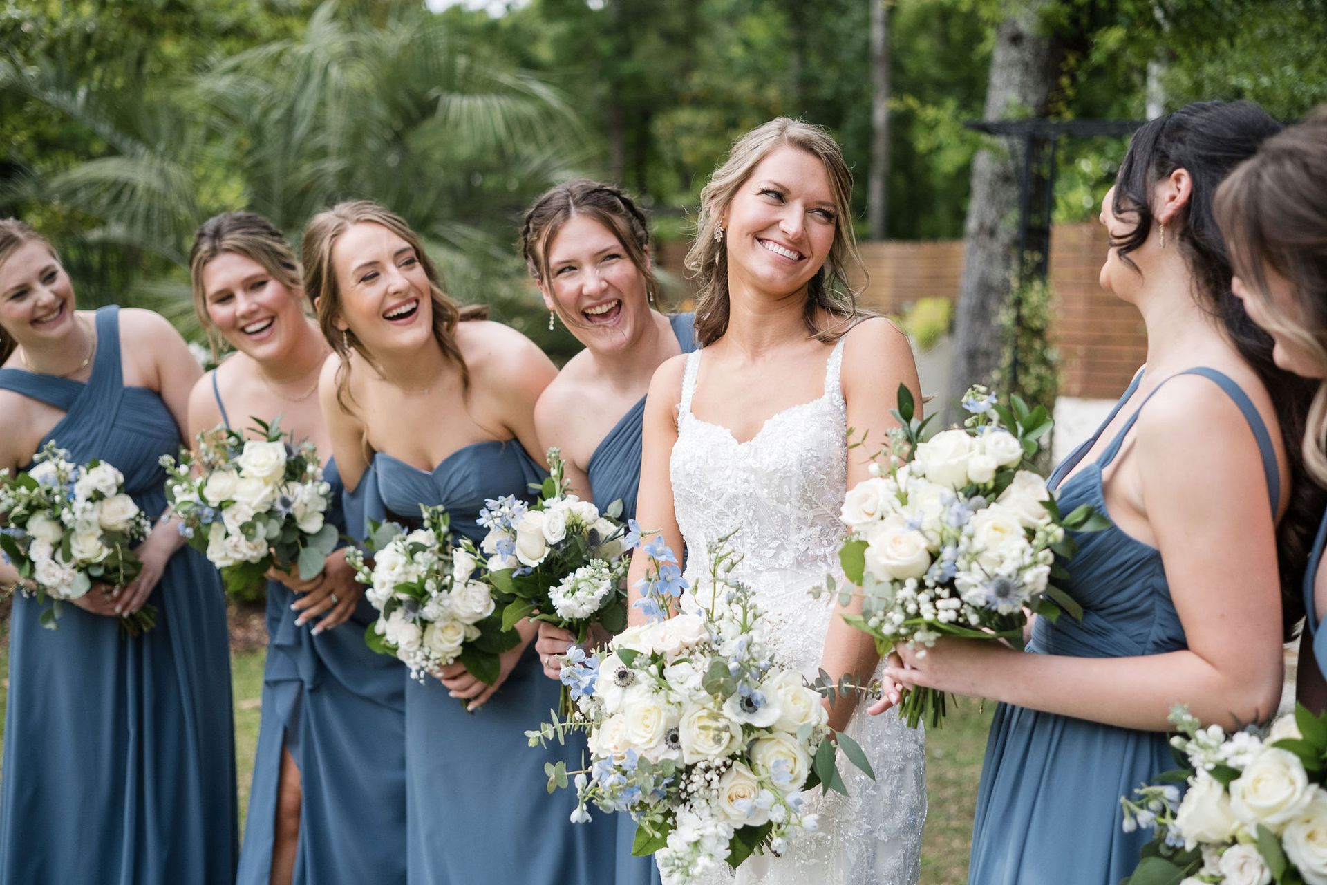Bridesmaids in blue dresses holding white bouquets, smiling outdoors at a wedding.