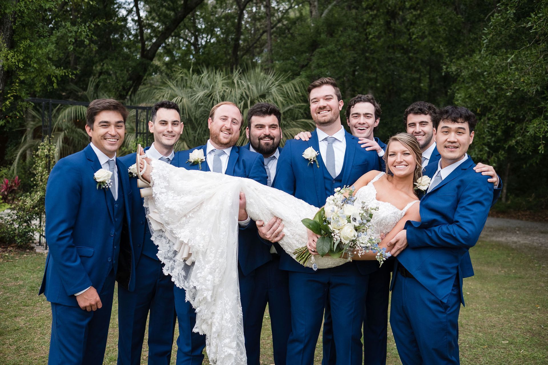 Wedding party in blue suits lifting the bride in a white dress outdoors among trees