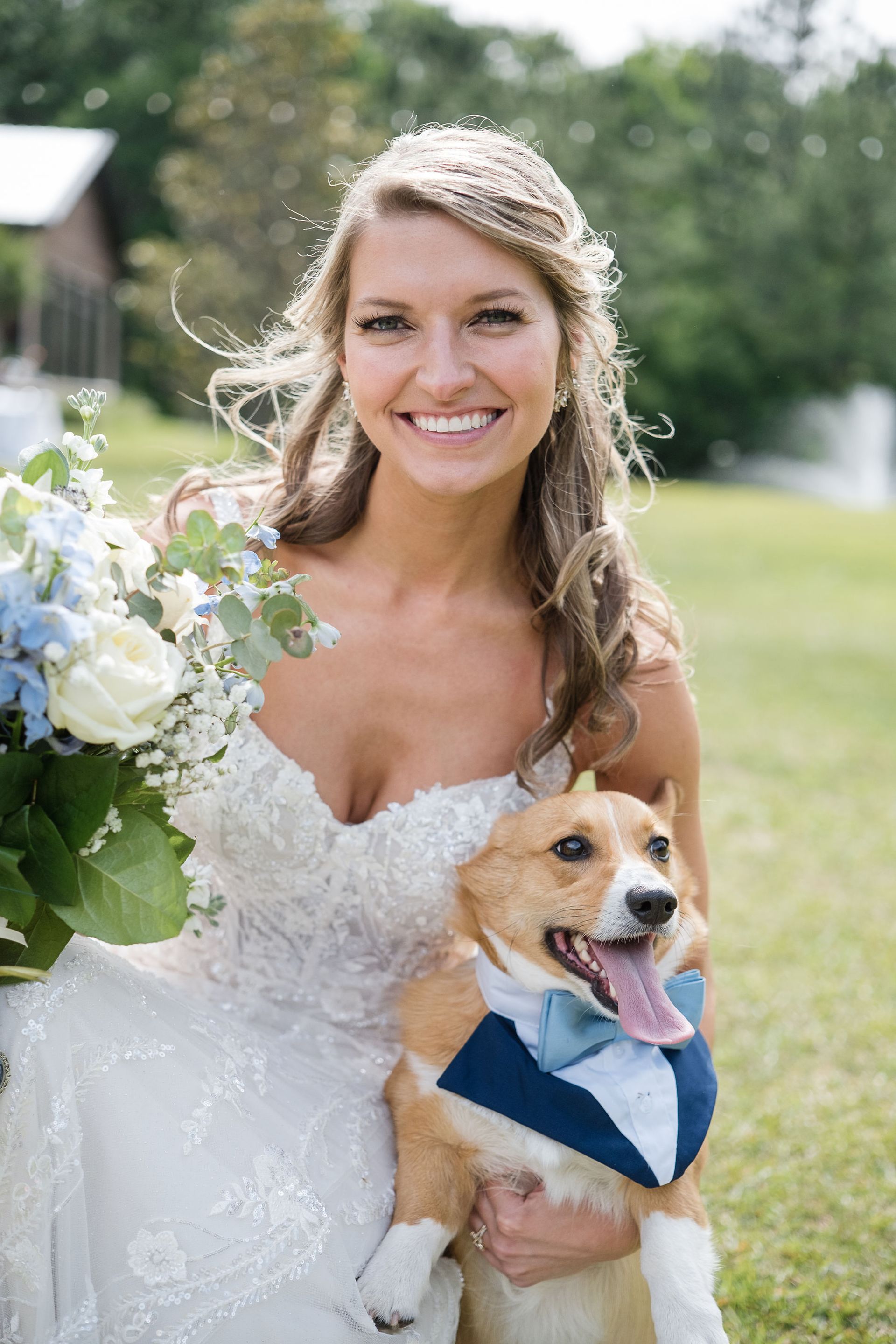 Bride in a white lace dress holding a bouquet and a small dog in a blue bandana outdoors