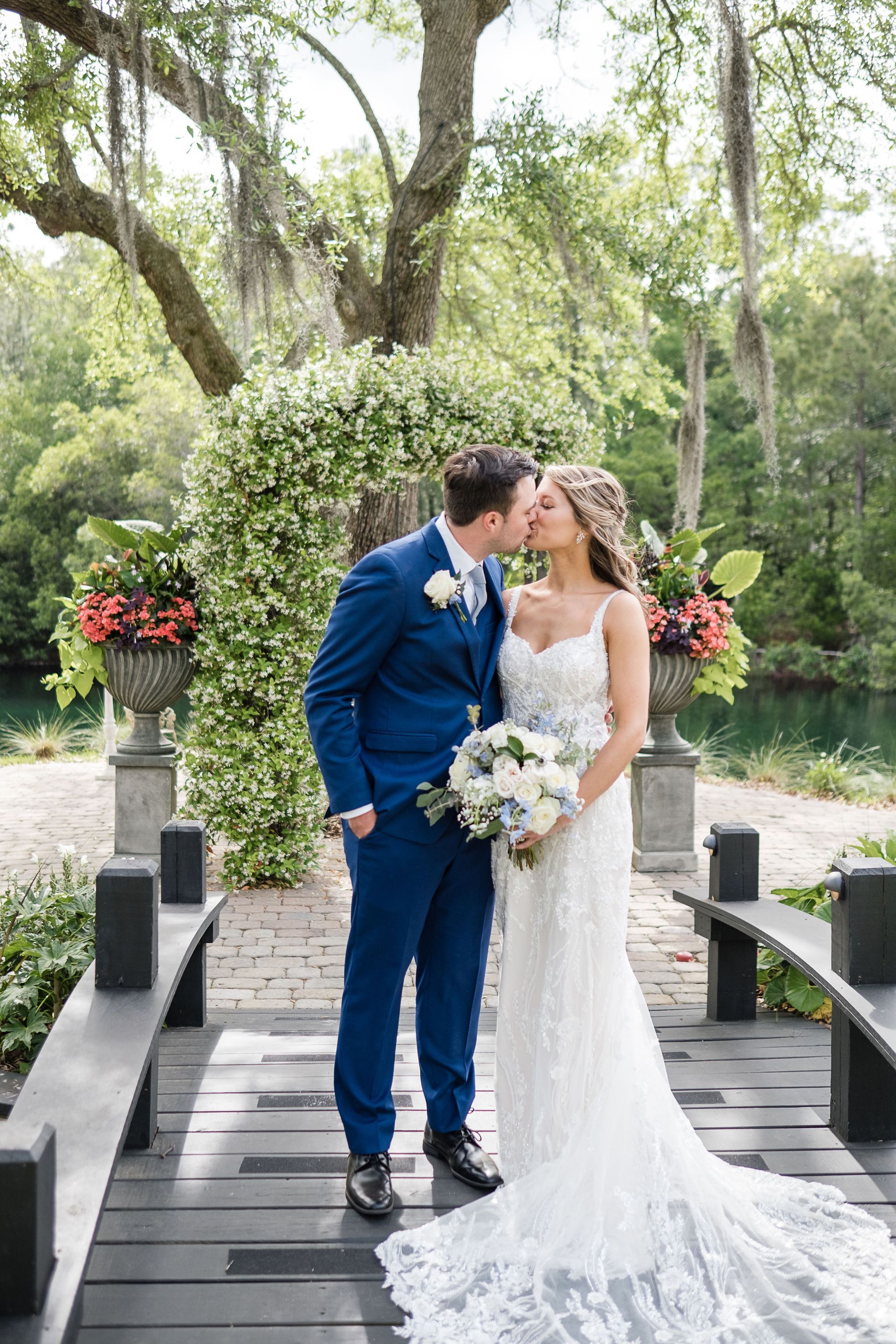 Bride and groom kissing on a stone bridge in a lush garden, holding a bouquet and wearing white and blue attire