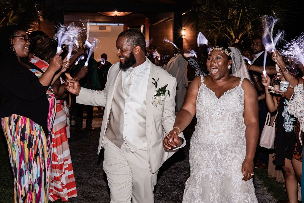 Newlyweds walk hand in hand through a sparkler send-off at night, both in white wedding attire.