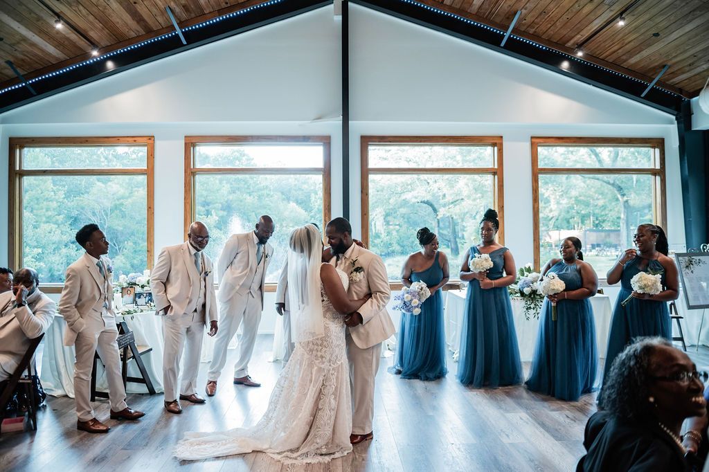 Wedding party standing in a bright hall, with bride and groom centered and guests lined along the windows