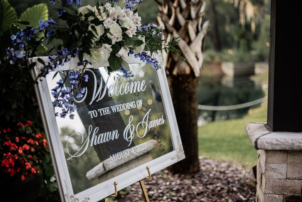 Wedding welcome sign with white flowers and greenery displayed outdoors beside a stone pillar.