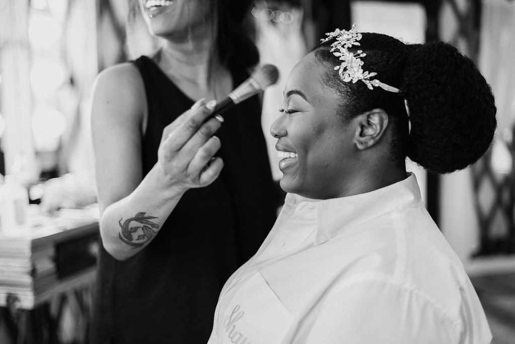 Person in a salon chair getting makeup applied, smiling as a stylist brushes their face.