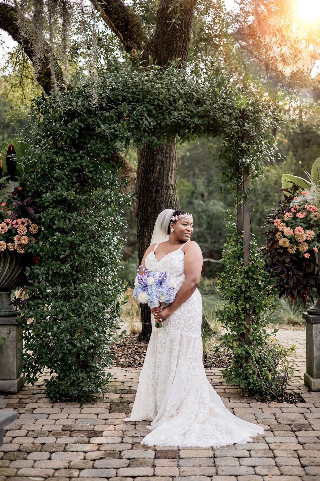 Bride in a white lace gown holding a blue bouquet under a floral arch in a garden courtyard