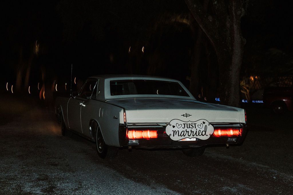 White classic car parked at night on a dark street, rear lights glowing.